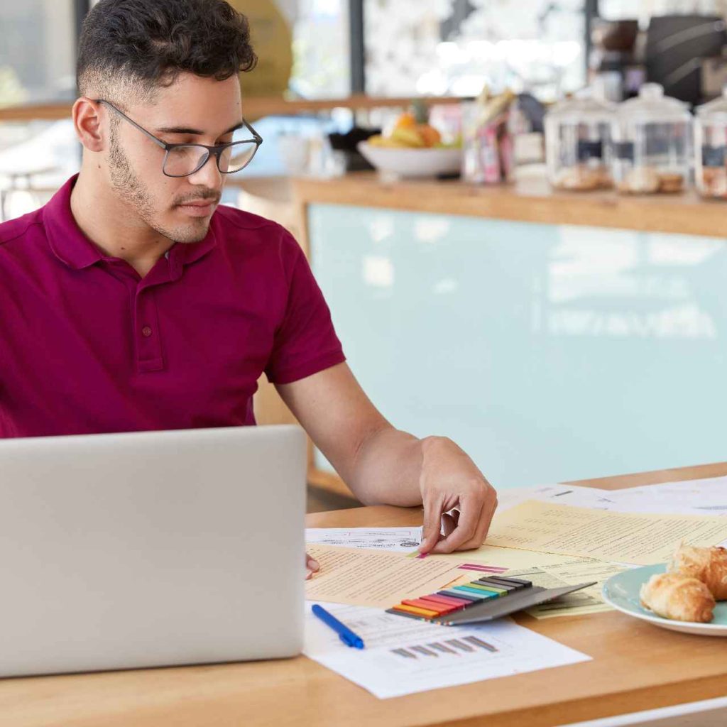 shot-young-male-uses-laptop-computer-checking-exchange-rate-internet-works-with-financial-documents-leaves-stickers-some-papers-going-have-tasty-lunch-cozy-snack-bar (1)
